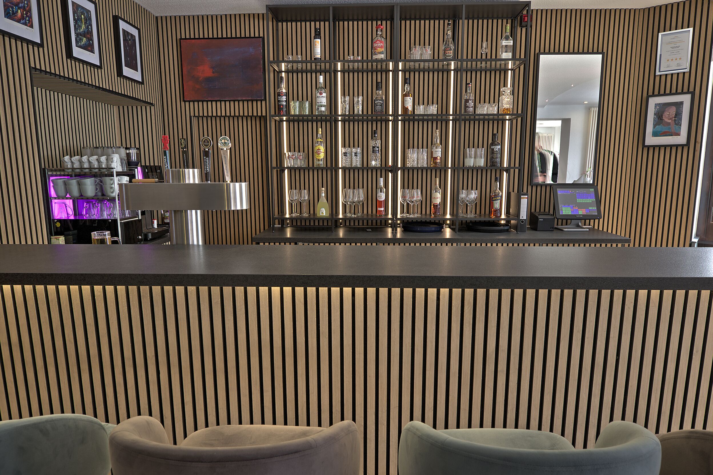 Un espace bar avec des étagères remplies de bouteilles et de verres, un comptoir et des sièges situés au premier plan.