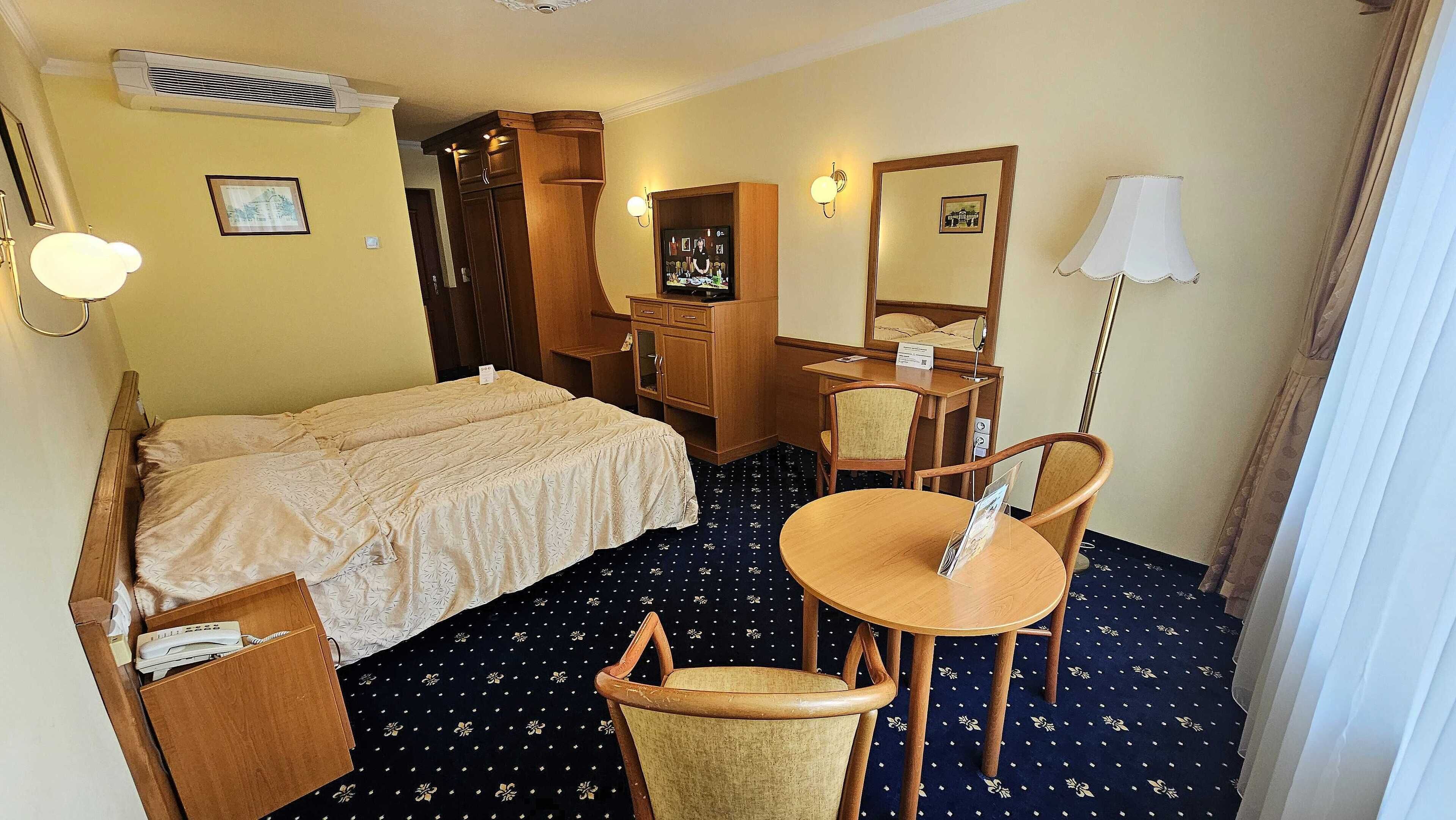 Habitación de hotel con cama, escritorio, silla, mesita, lámpara y equipo de aire acondicionado.