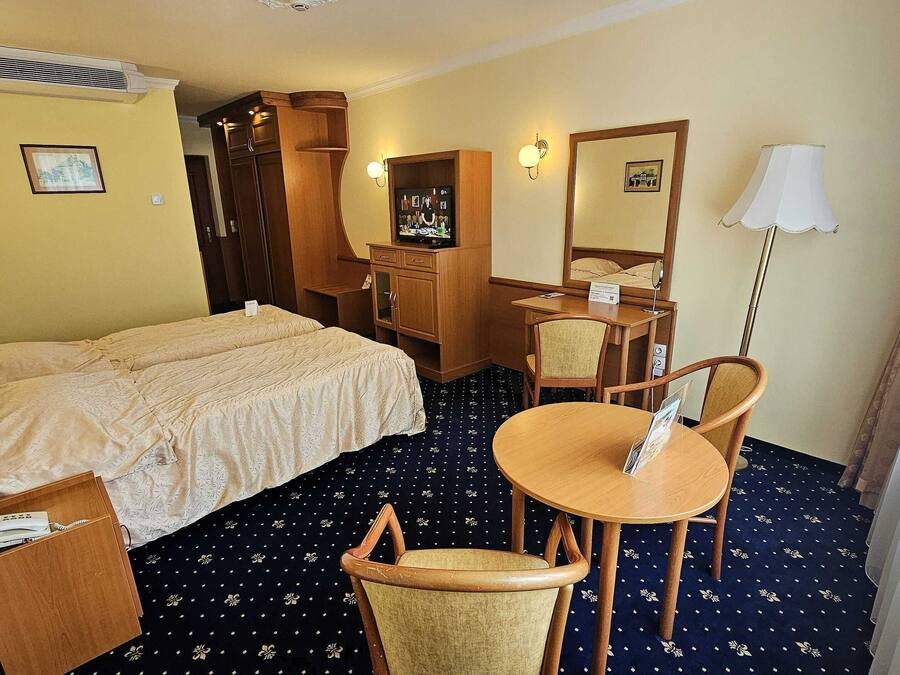 A hotel room with a bed, a desk, a chair, a small table, a lamp, and an air conditioning unit.