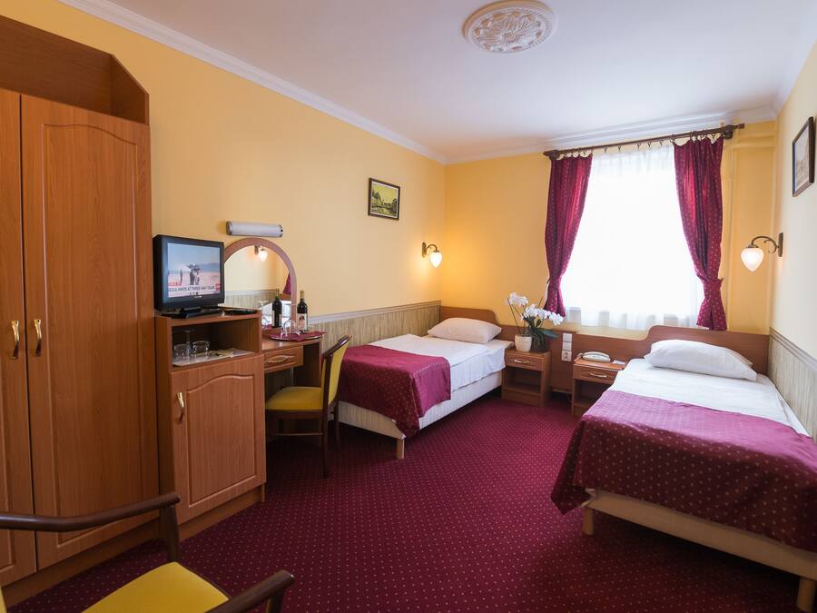 Habitación de hotel con dos camas individuales, un escritorio con ordenador, una silla, un armario y una ventana con cortinas.