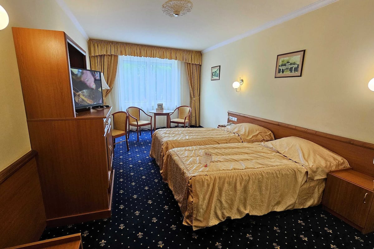Ein Hotelzimmer mit zwei Betten, einem Schreibtisch mit Stühlen, einem Fernseher und einem Fenster mit Vorhängen.
