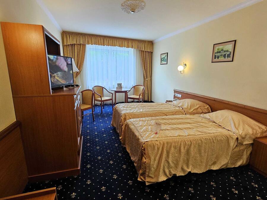 A hotel room with two beds, a desk with chairs, a television, and a window with curtains.