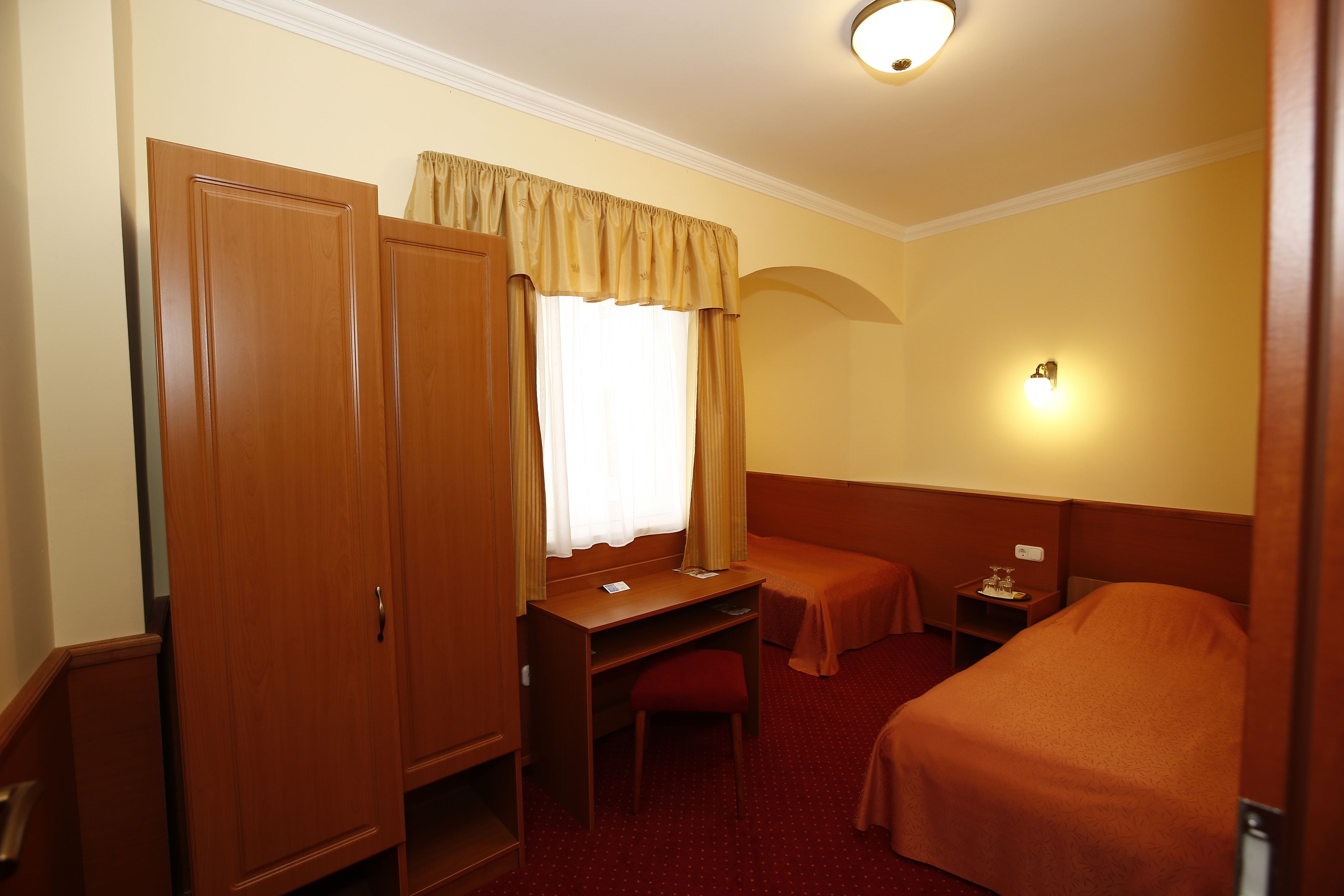 A hotel room with a wooden desk, a single bed, a small table with a lamp, and a wardrobe.