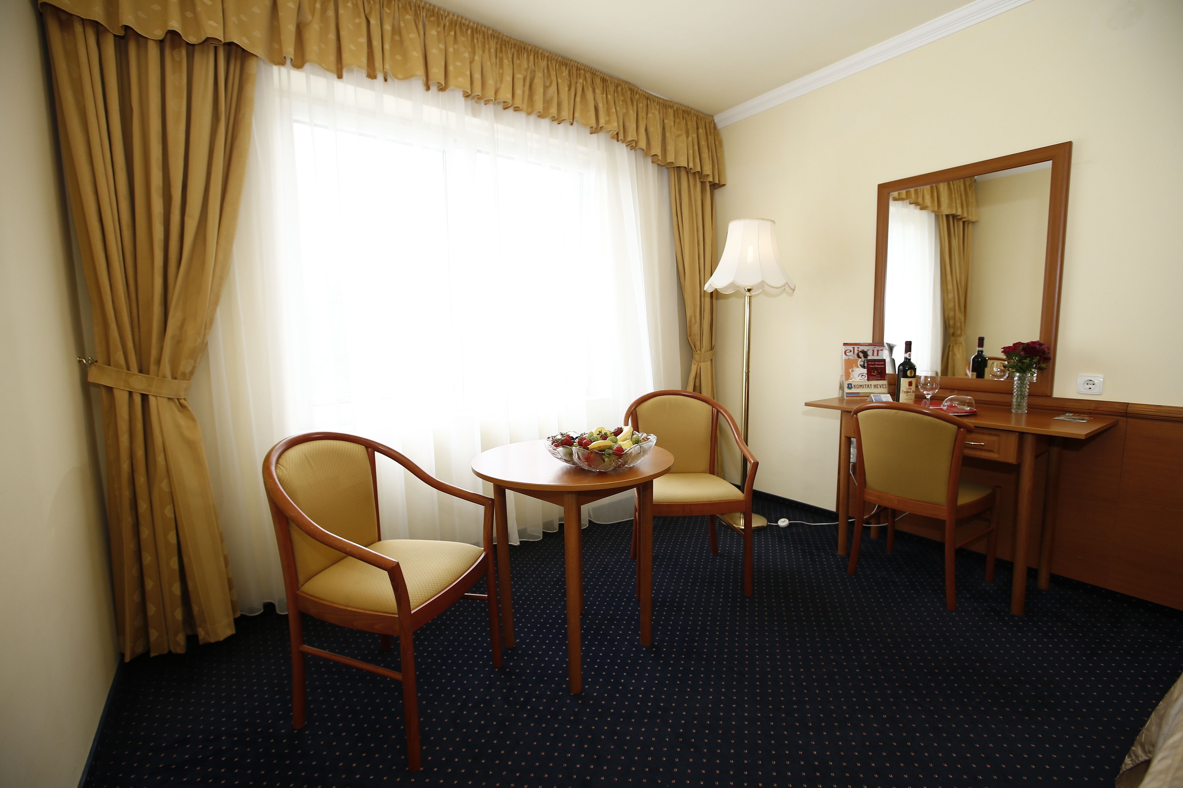 A hotel room with two wooden chairs, a small round table with a fruit bowl, a desk with a lamp, and a mirror.