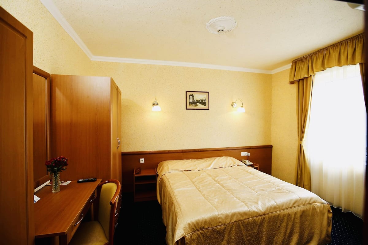Ein Hotelzimmer mit Bett, Schreibtisch mit Blumenvase, Stuhl, Nachttisch und einem Bild an der Wand.