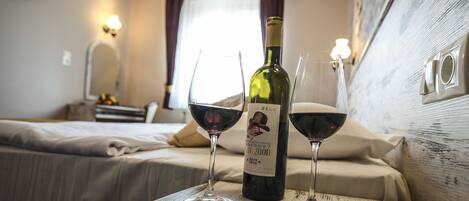 Habitación de hotel con cama, una mesa de madera con una botella de vino y dos copas de vino tinto, y una lámpara de pared.
