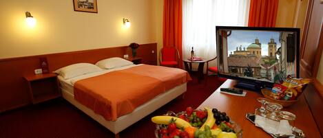 A hotel room with a bed, a desk with a fruit bowl, a television, and a window with curtains.