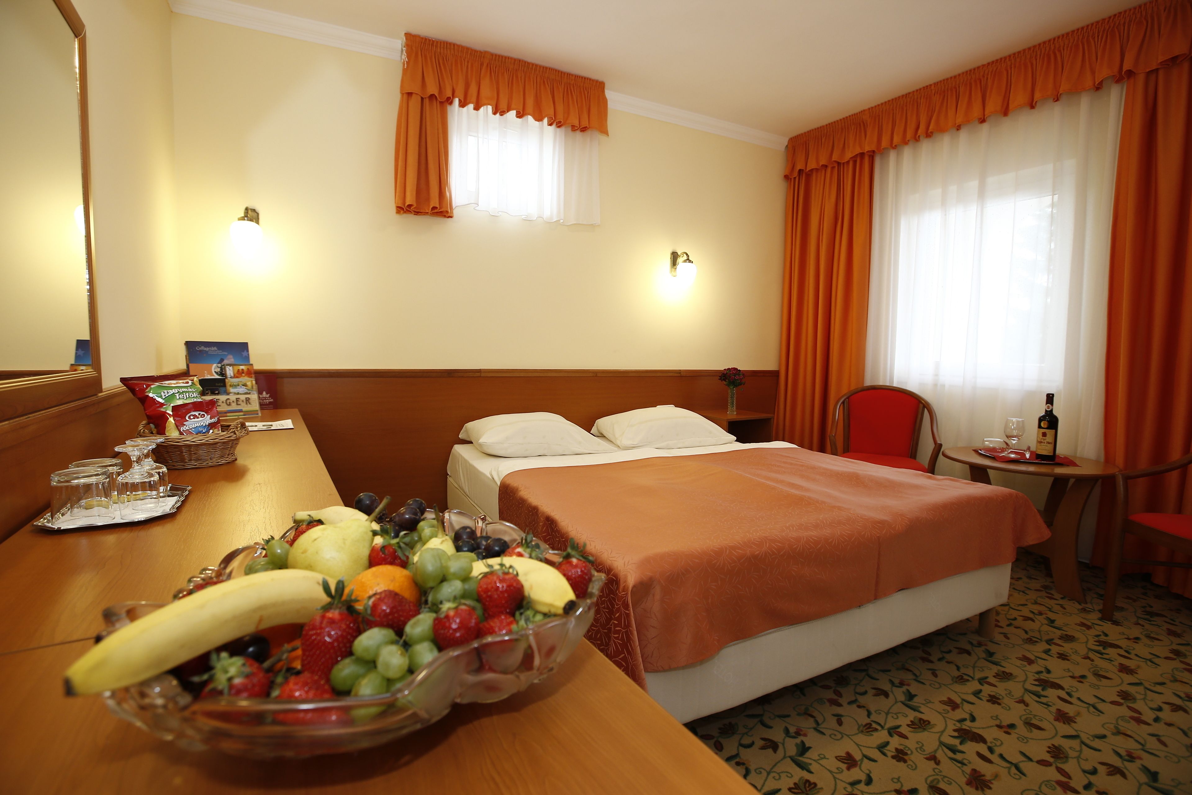 Habitación de hotel con cama, una mesa de comedor con fruta, una silla y una mesita con una botella y un vaso.