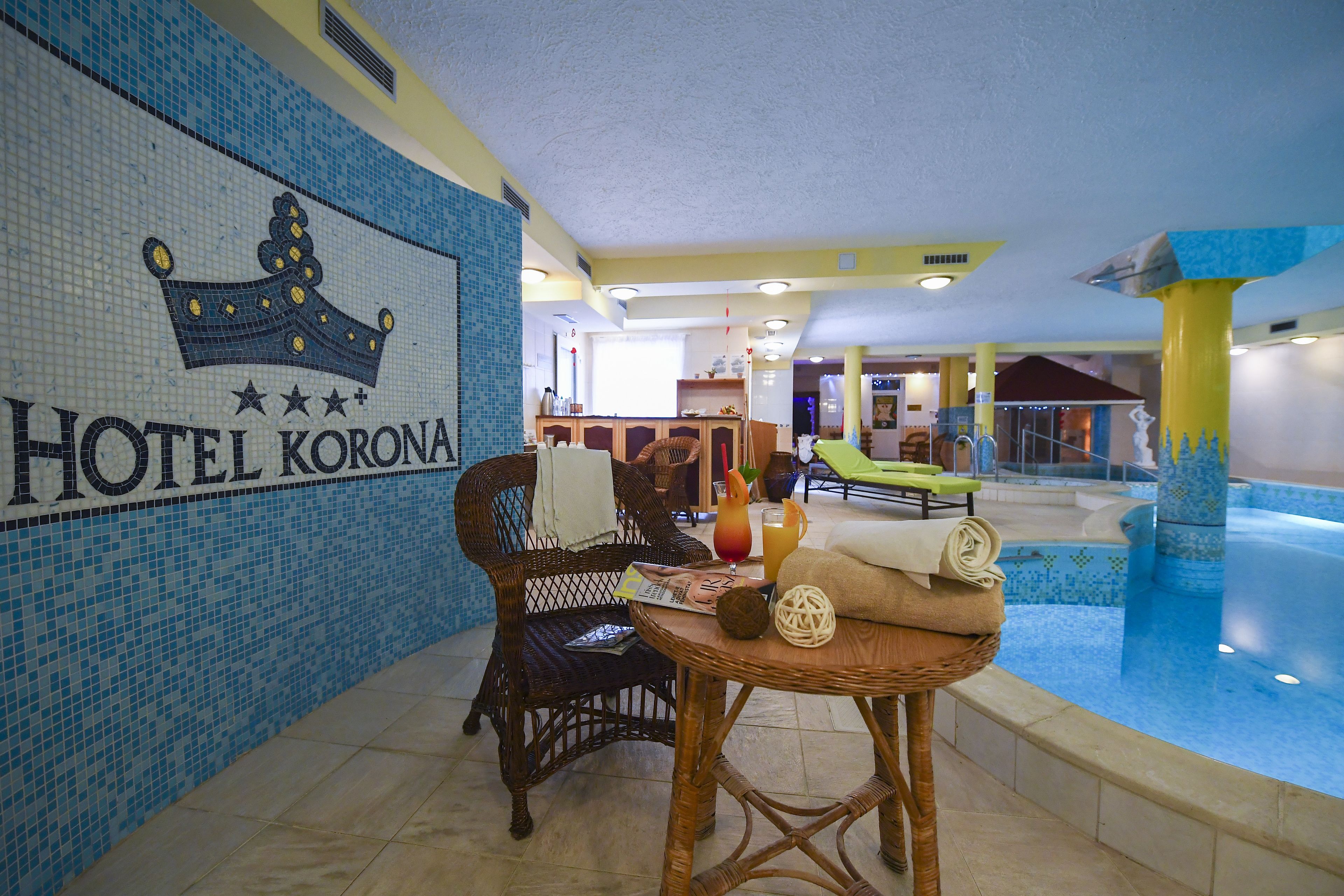 A hotel lobby with a tiled wall, a wicker chair, a small table with towels and a fruit bowl, and a pool area in the background.