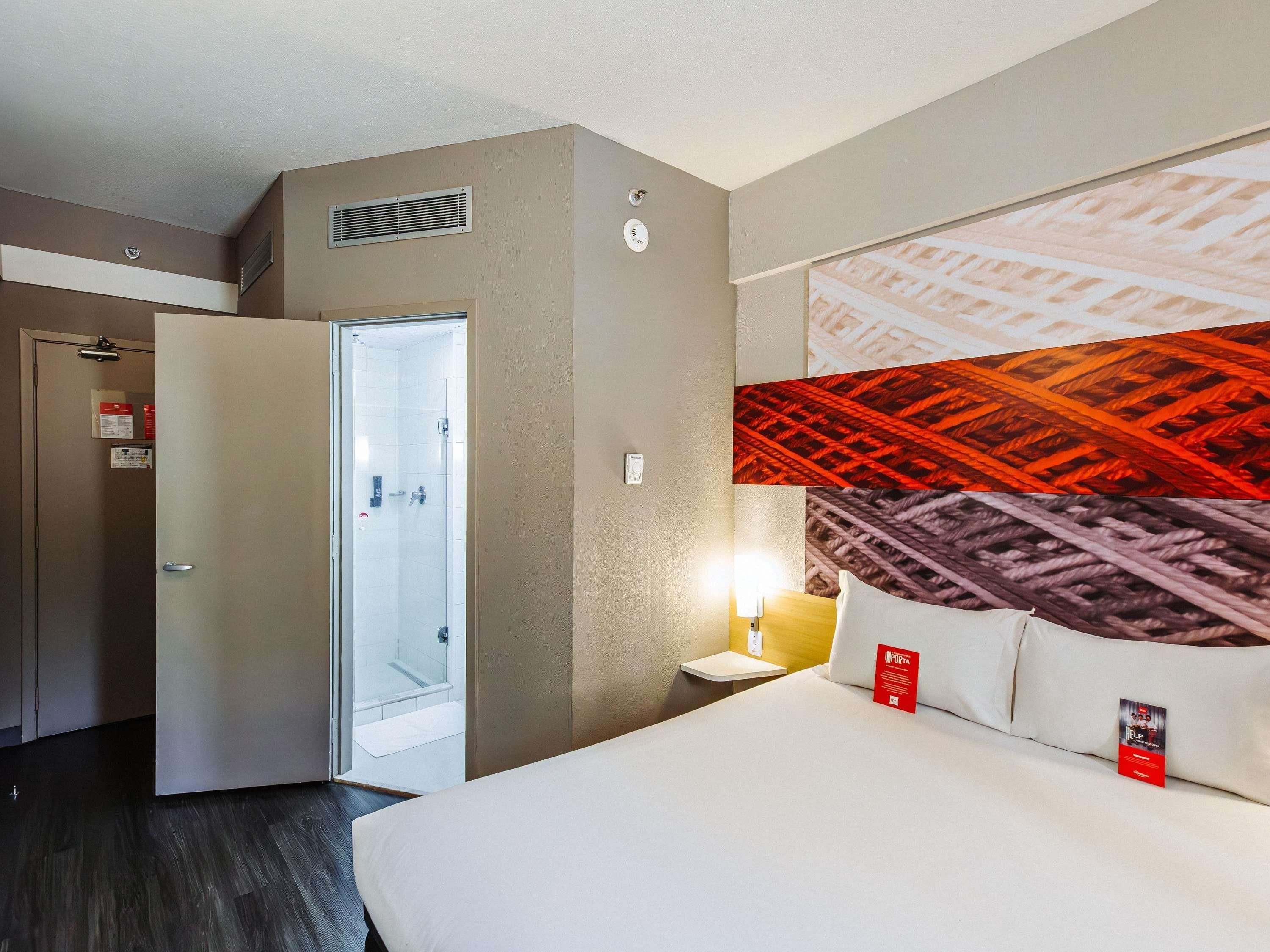 A hotel room with a bed, a nightstand, a wall-mounted lamp, a door leading to a bathroom, and a large abstract wall art piece.