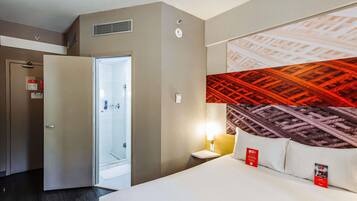 A hotel room with a bed, a nightstand, a wall-mounted lamp, a door leading to a bathroom, and a large abstract wall art piece.