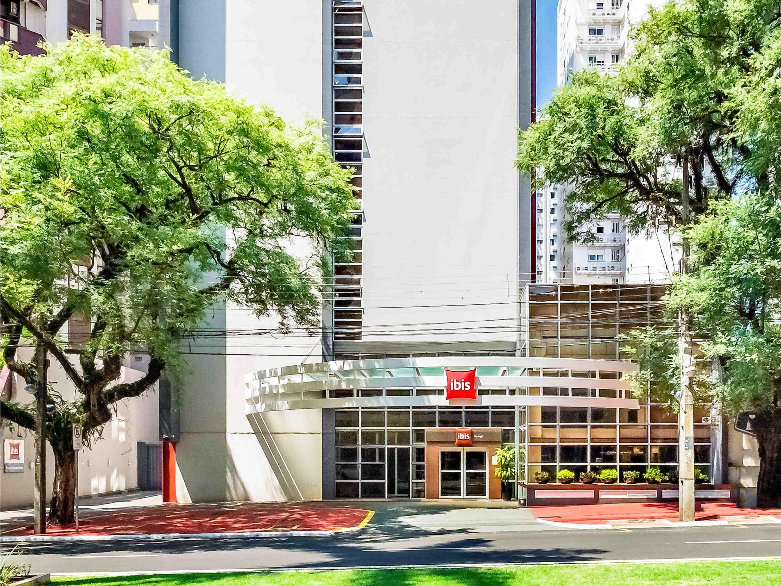 A modern hotel with a glass facade, a red "ibis" sign, and a landscaped entrance area.