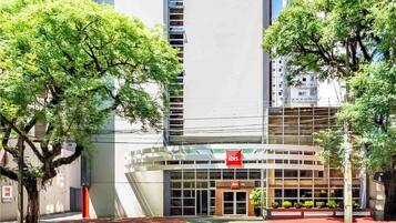 A modern hotel with a glass facade, a red "ibis" sign, and a landscaped entrance area.