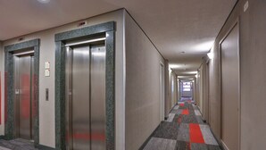 Un pasillo de hotel con dos ascensores, pisos alfombrados y un largo corredor que conduce a una puerta al final.