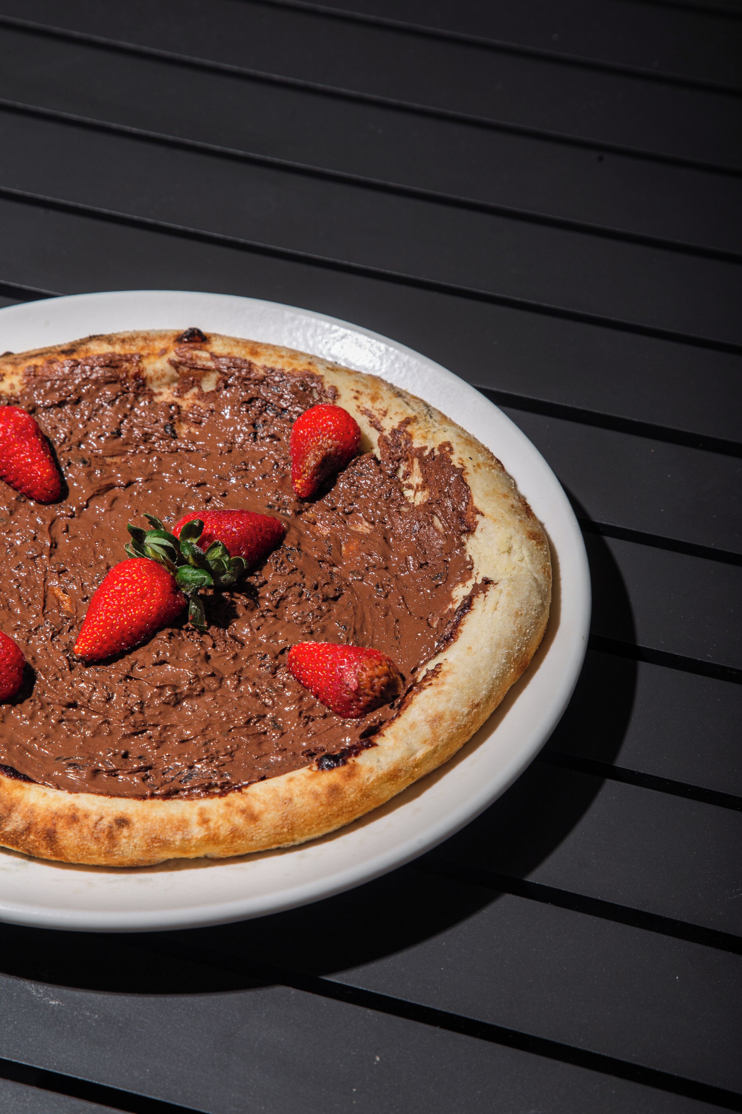 Eine mit Schokolade überzogene Pizza auf einem weißen Teller, dazu Erdbeeren auf dunklem Untergrund.