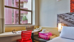 Habitación de hotel con escritorio, una silla roja, una cama, una maleta y vistas a un edificio a través de una ventana.