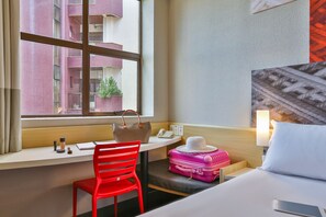 A hotel room with a desk, a red chair, a bed, a suitcase, and a view of a building through a window. - ibis Maringa (Maringá)