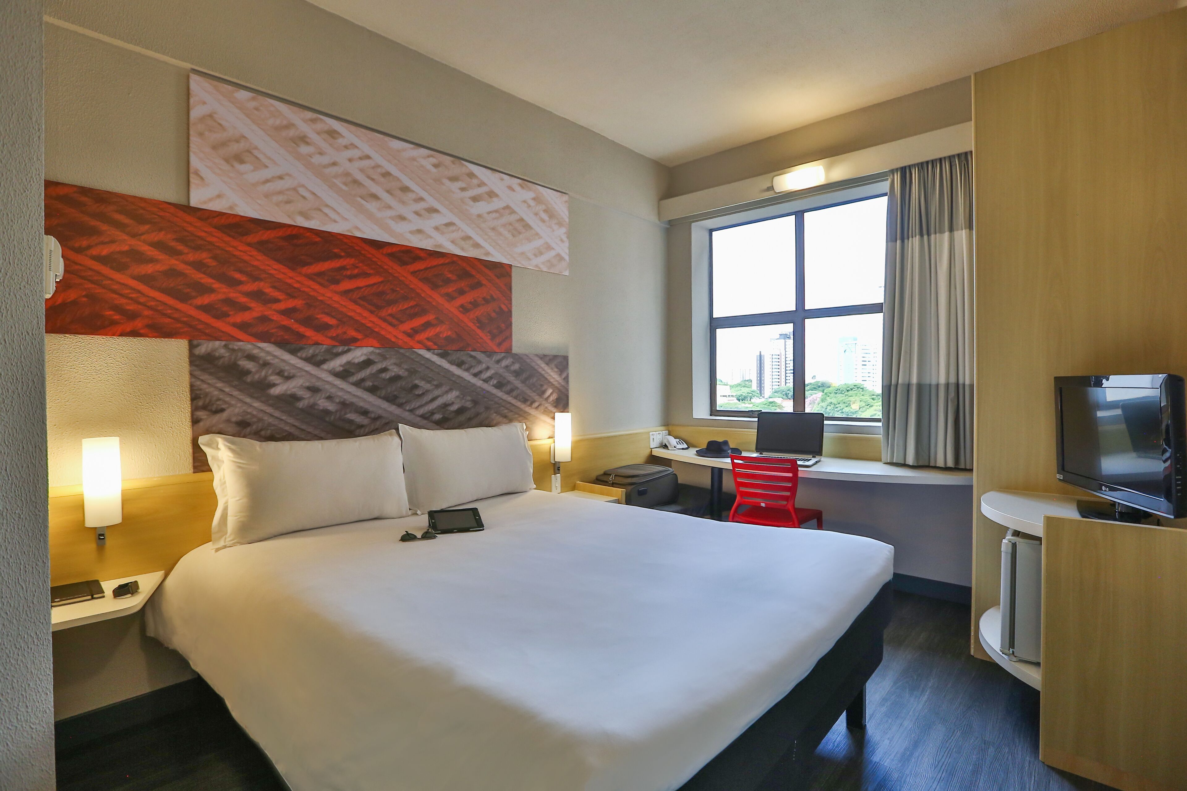 Habitación de hotel con una cama grande, un escritorio con computadora, televisión y ventana con vista a edificios.