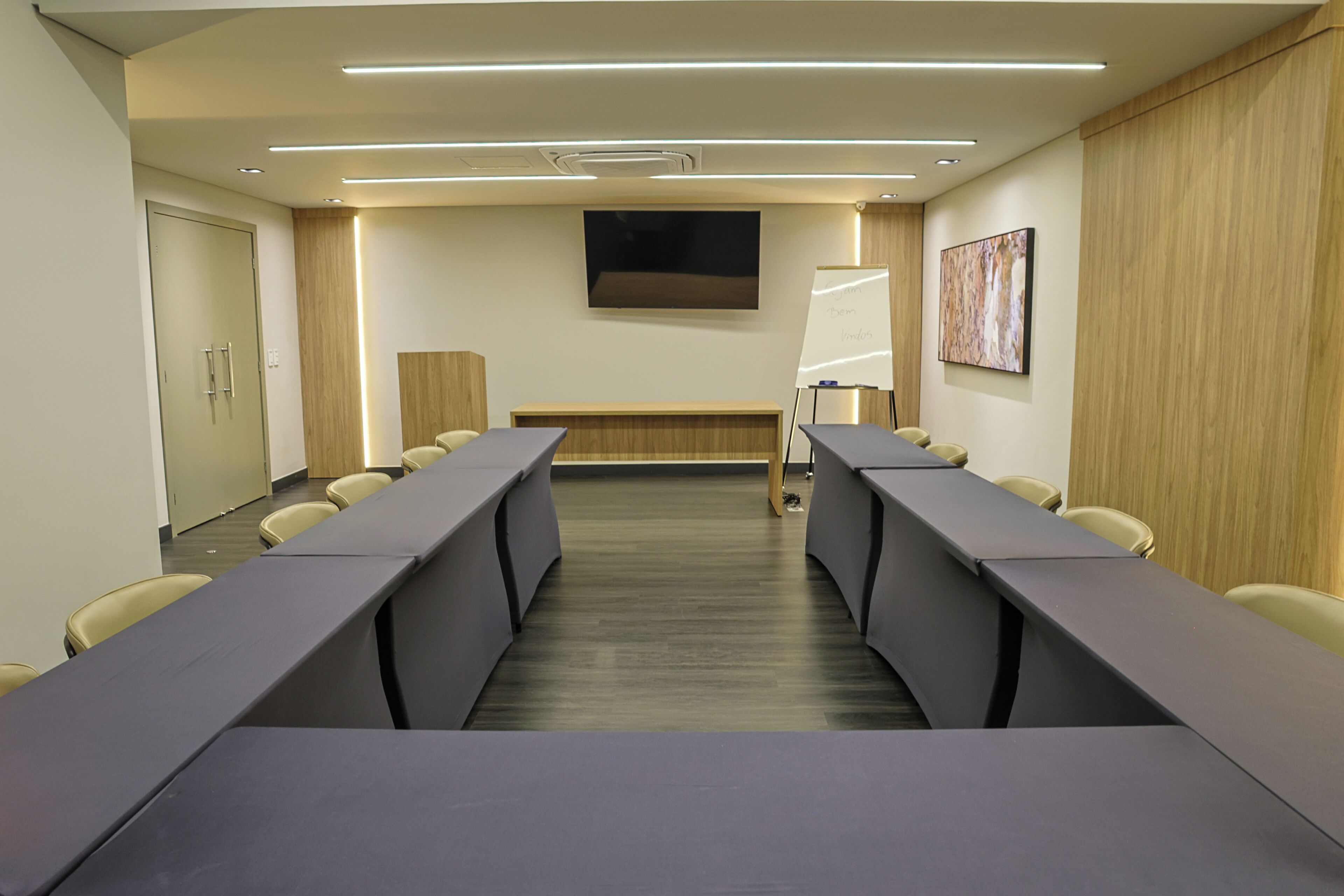 Uma sala de conferências com uma mesa comprida, cadeiras, um quadro branco e uma televisão na parede.