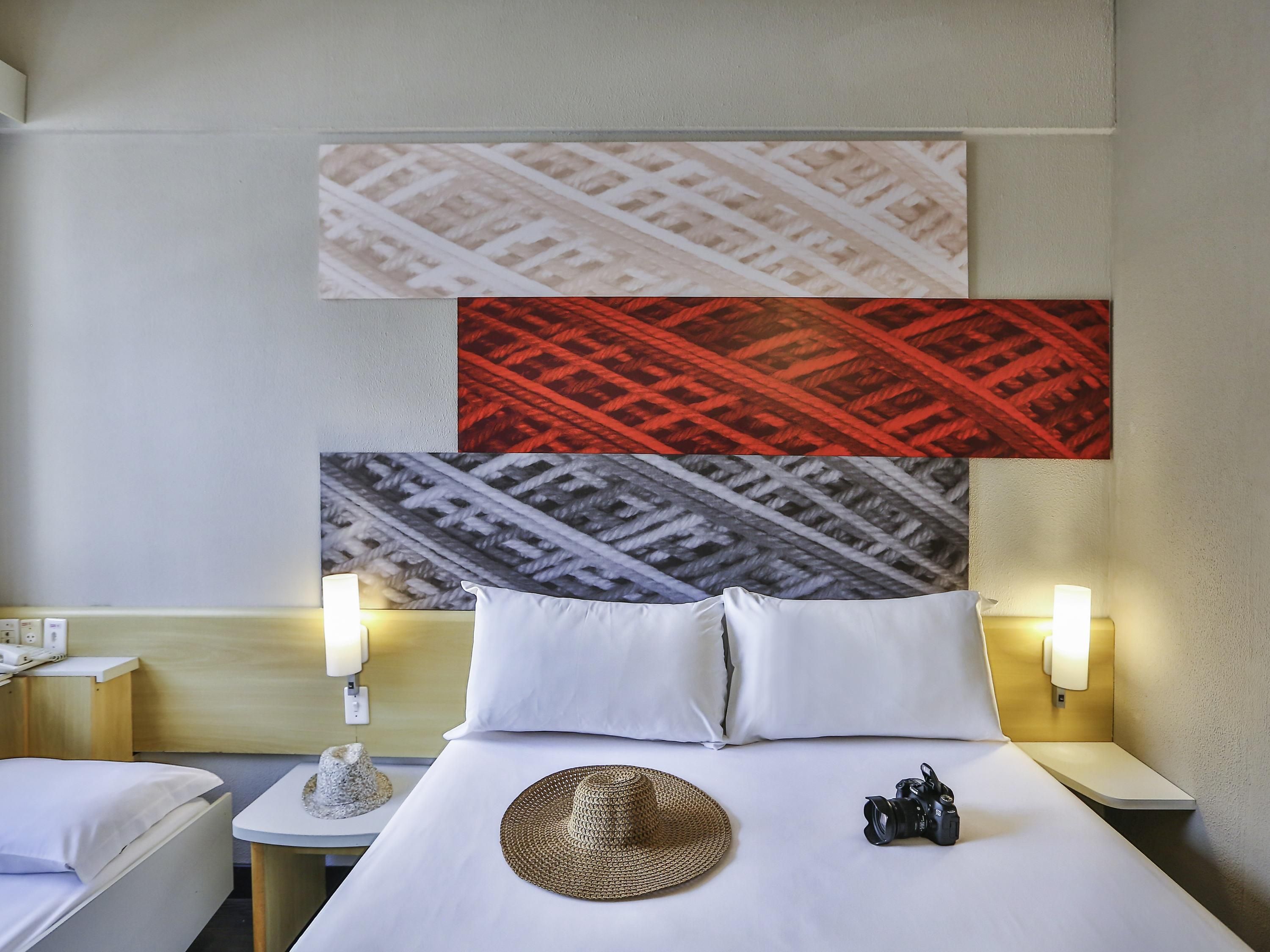 Habitación de hotel con cama, dos lámparas de noche, una obra de arte en la pared, un sombrero y una cámara sobre la cama.