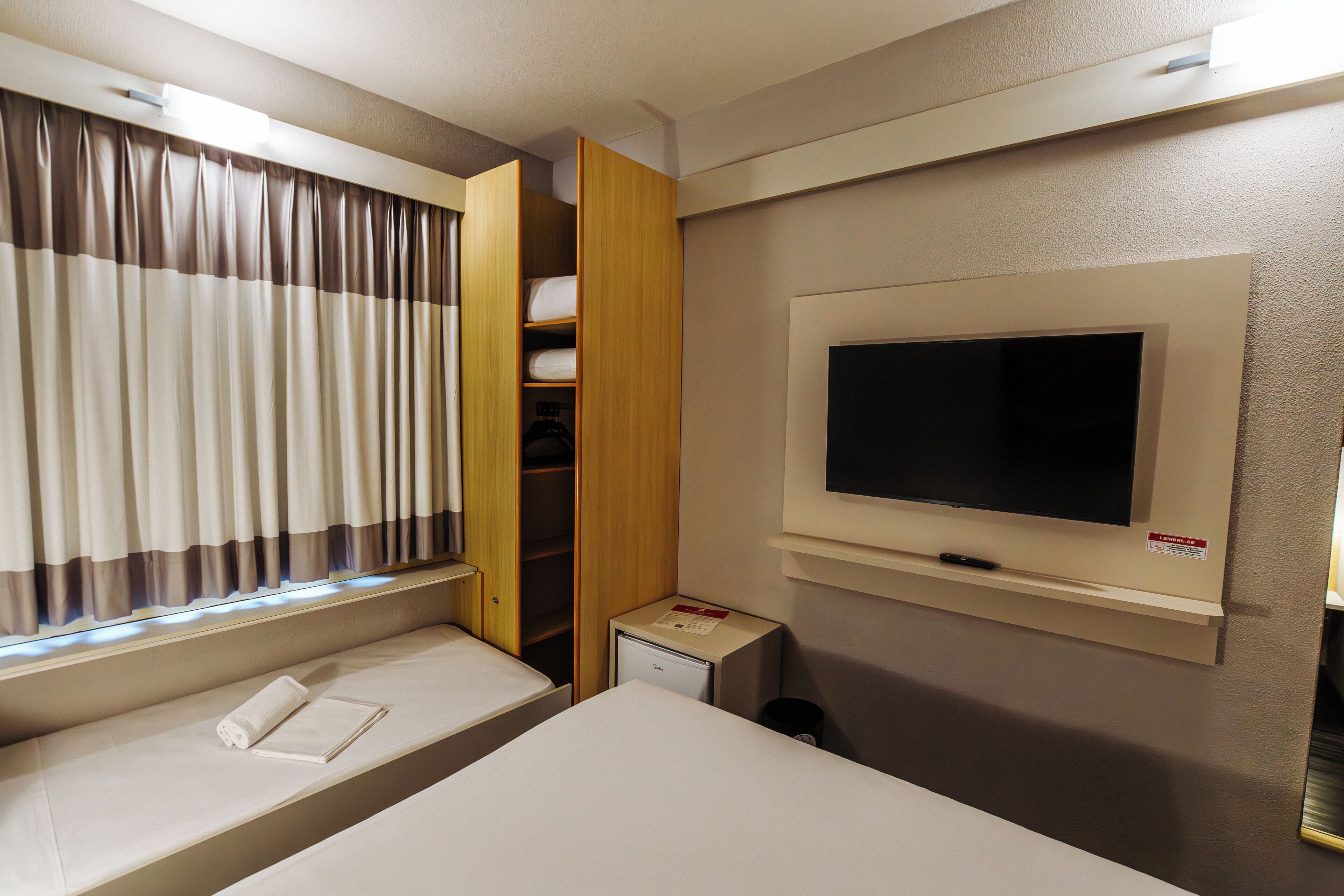 A hotel room with a bed, a TV mounted on the wall, a window with curtains, and a built-in wardrobe.