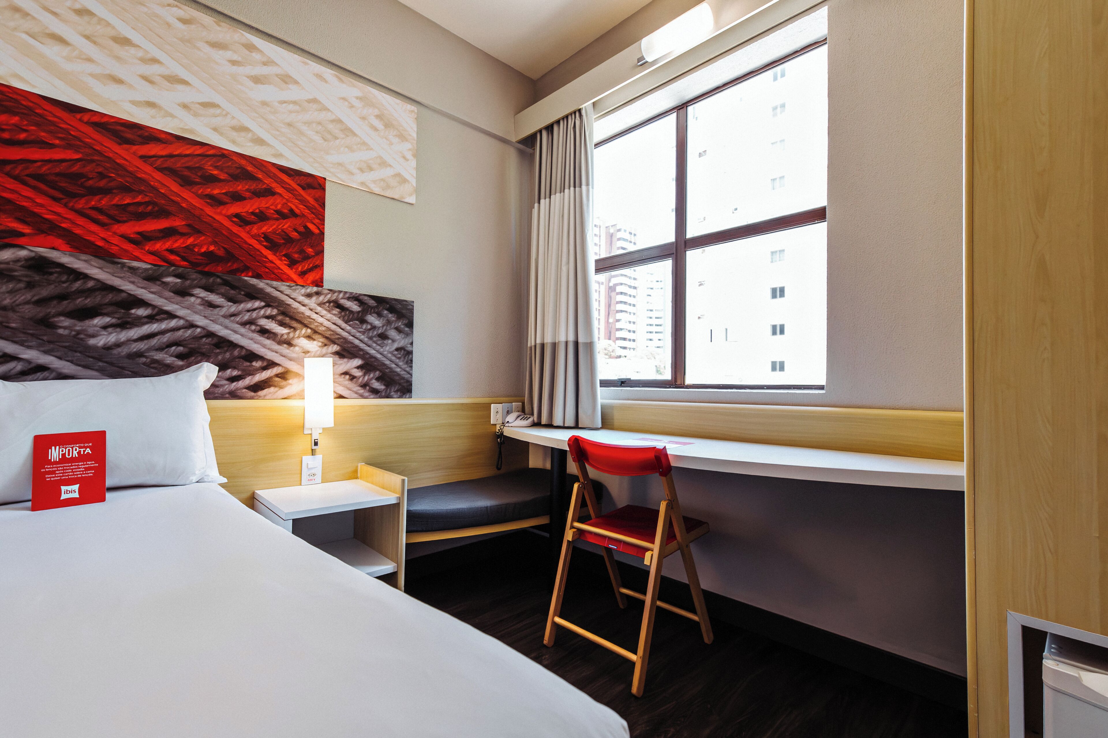 Een hotelkamer met een bed, een bureau, een stoel, een raam met uitzicht op gebouwen, en een rode wanddecoratie.
