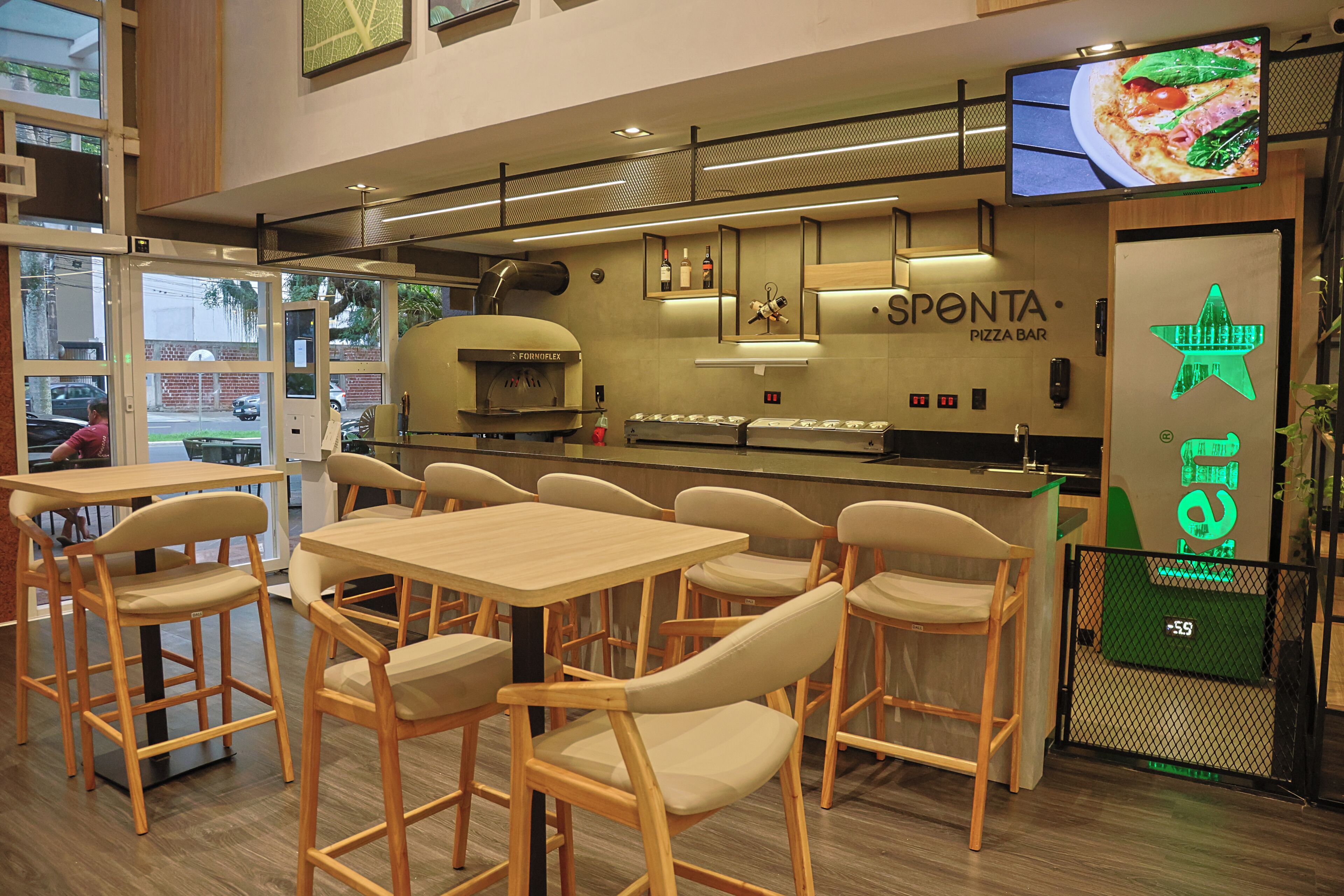 Um pizzabar com mesas e cadeiras de madeira, um forno a pizza e uma televisão exibindo pizzas.