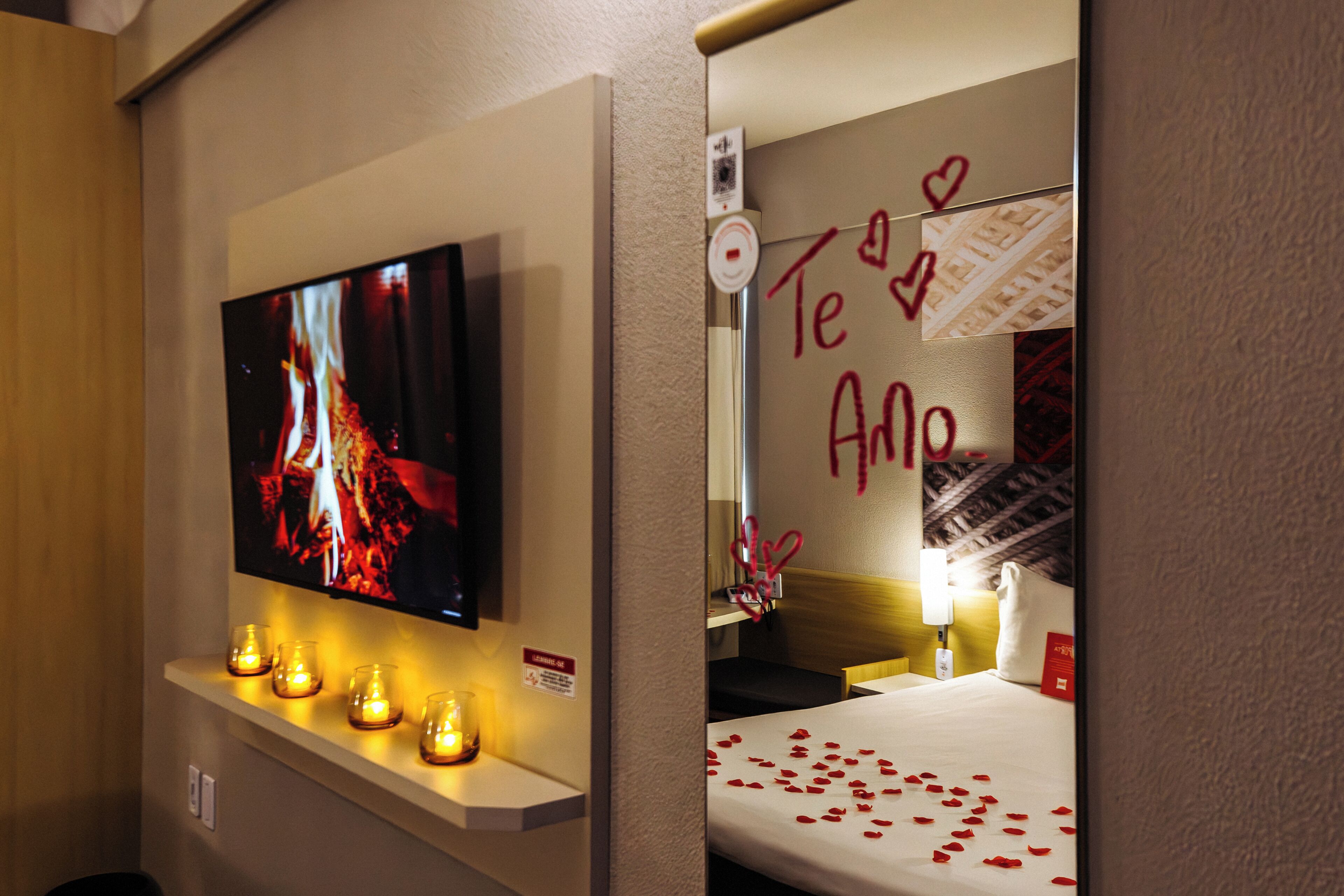 Quarto de hotel com televisão na parede, mesa de cabeceira com velas e cama decorada com pétalas de rosa.