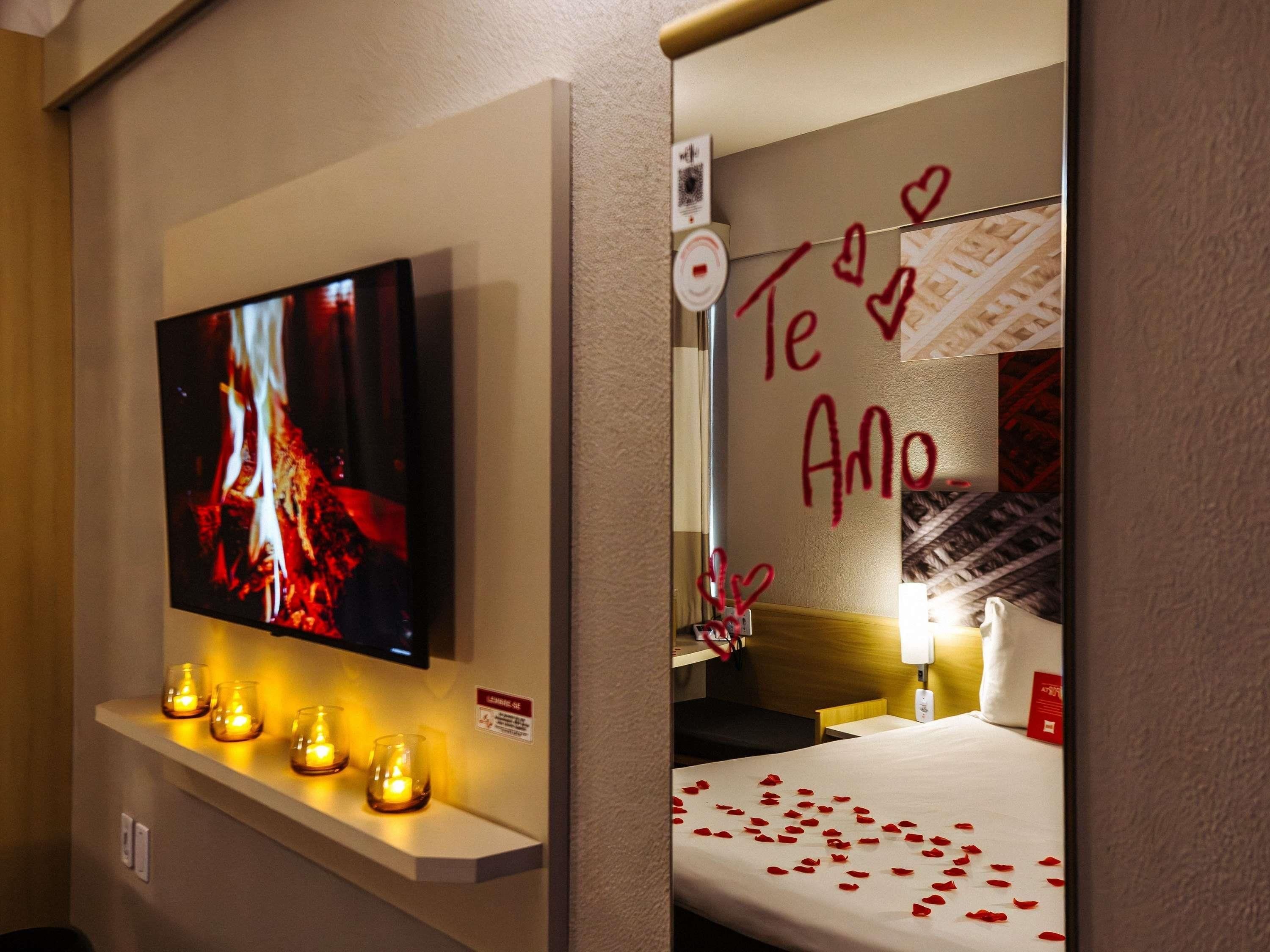 Une chambre d’hôtel avec un téléviseur fixé au mur, une étagère avec des bougies, un lit parsemé de pétales de rose et un mur orné d’une décoration en forme de cœur.