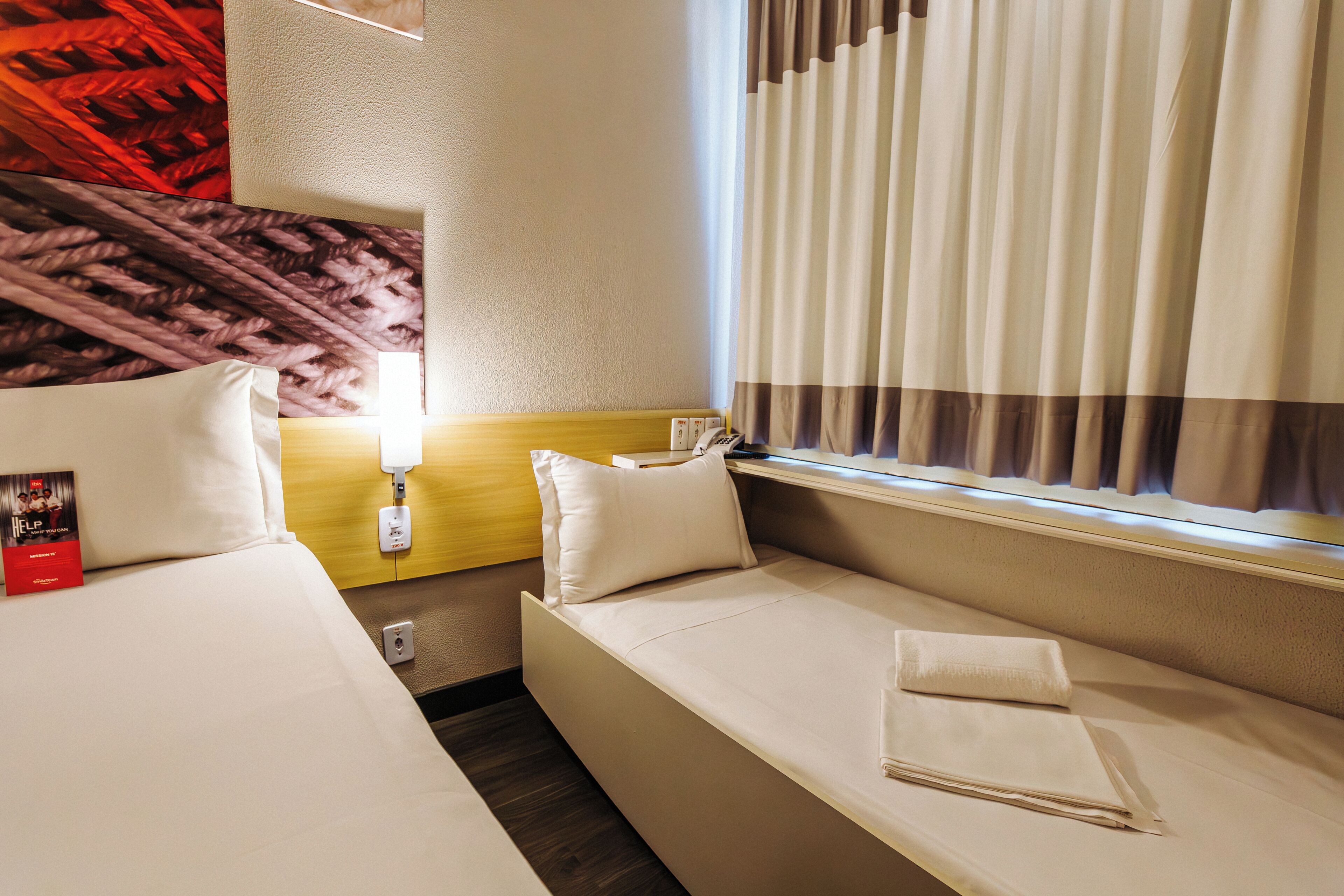 A hotel room with two beds, a small seating area, a window with curtains, and a wall-mounted telephone.