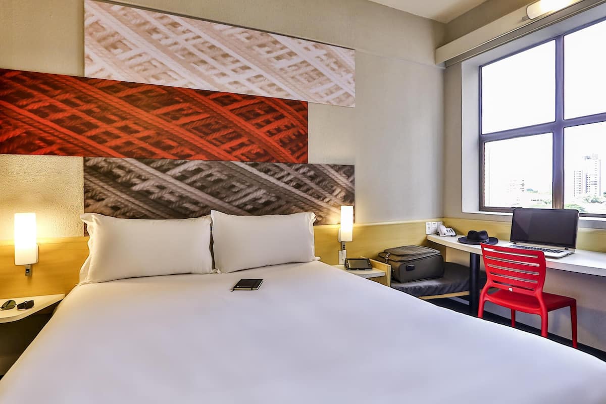 A hotel room with a bed, bedside lamps, a desk with a laptop, a red chair, and a window with a city view.