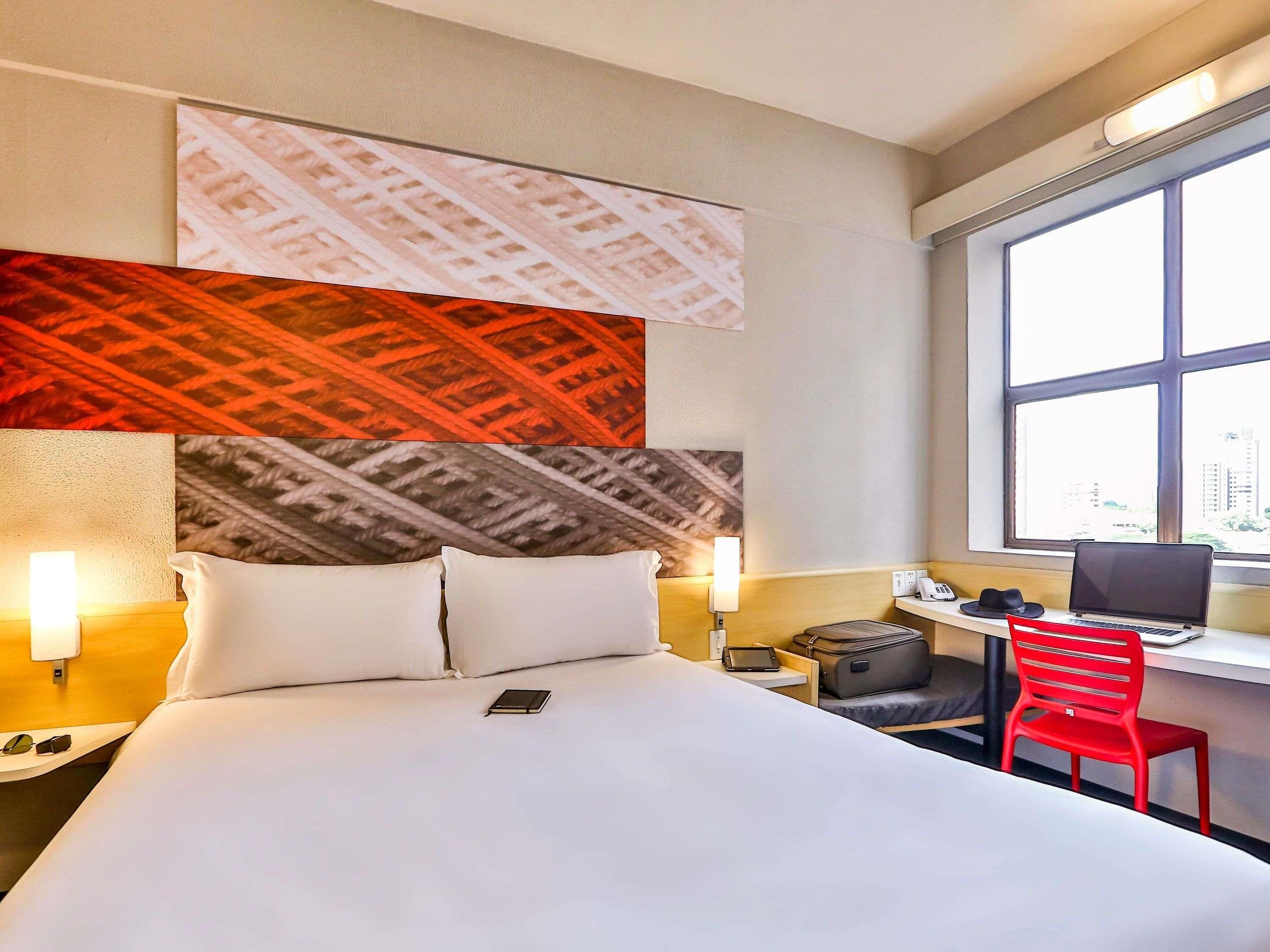 A hotel room with a bed, bedside lamps, a desk with a laptop, a red chair, and a window with a city view.
