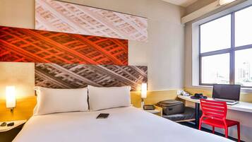 A hotel room with a bed, bedside lamps, a desk with a laptop, a red chair, and a window with a city view.