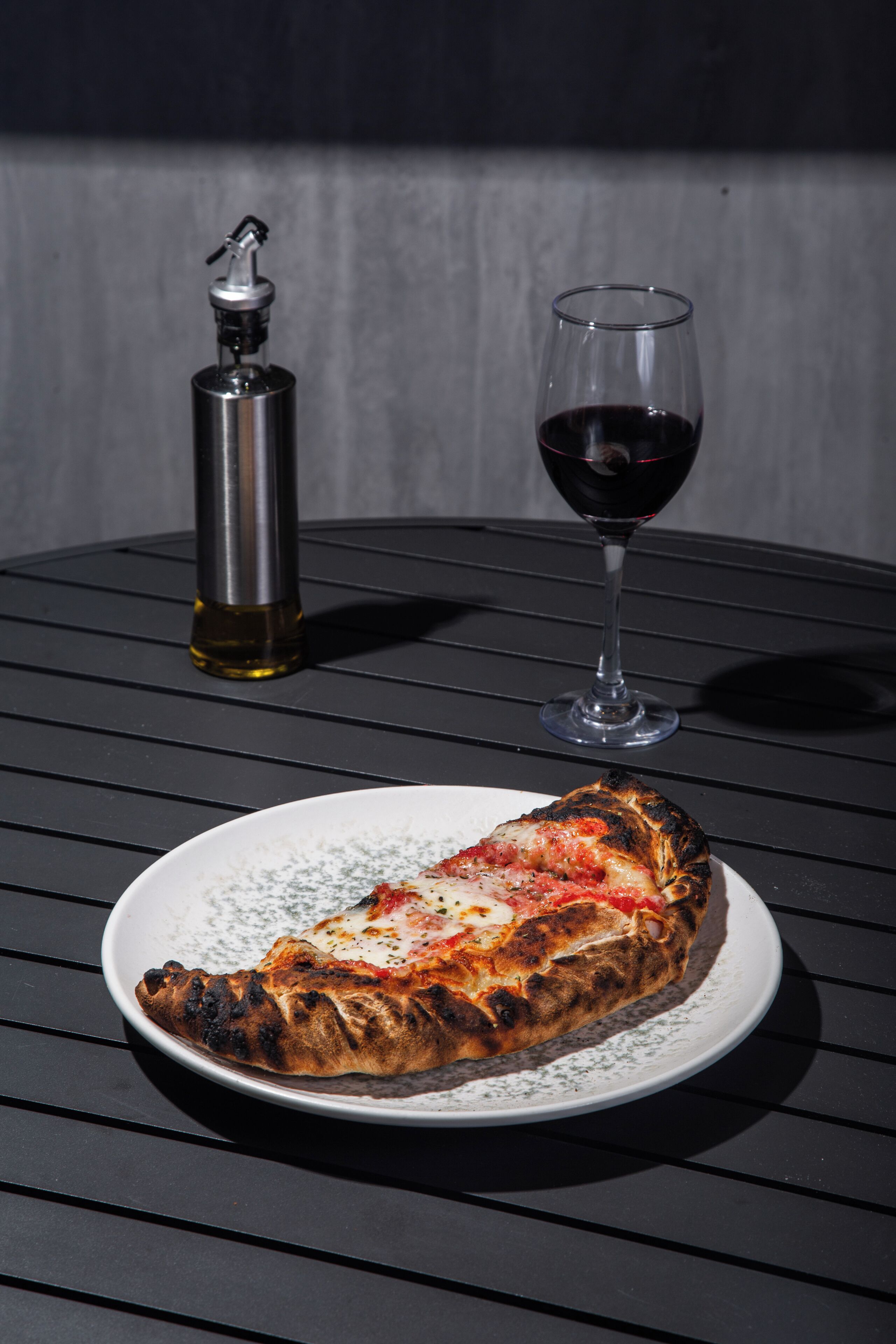 A single slice of pizza on a plate with a glass of red wine and a bottle of olive oil on a metal table.