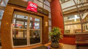 Exterior de un hotel con un llamativo letrero 'ibis', una entrada de vidrio y una planta en maceta al frente.