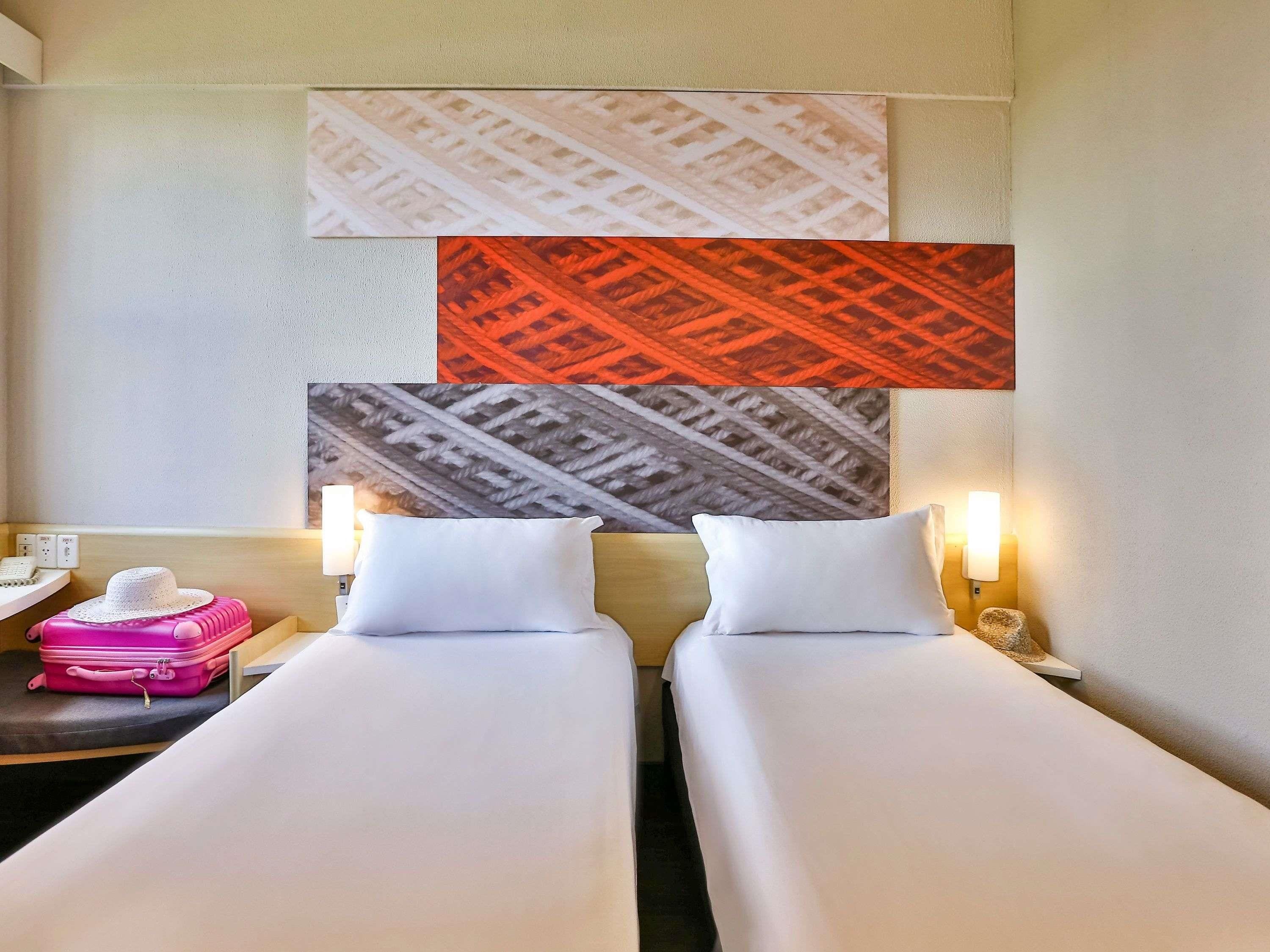 A hotel room with two beds, a nightstand with a lamp, a pink suitcase, and a wall with patterned artwork.