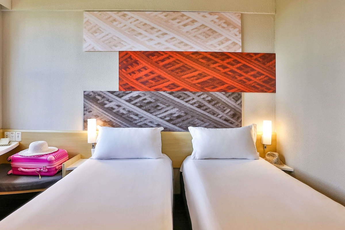 A hotel room with two beds, a nightstand with a lamp, a pink suitcase, and a wall with patterned artwork.