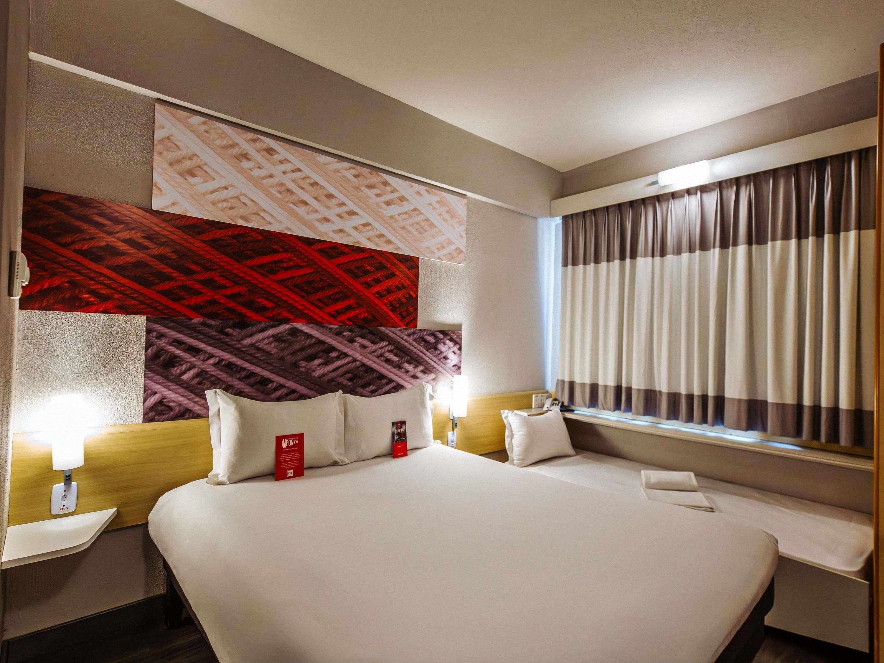 Quarto de hotel com uma cama grande, cabeceira com padrão decorativo, janela com cortinas e luminária fixada na parede.