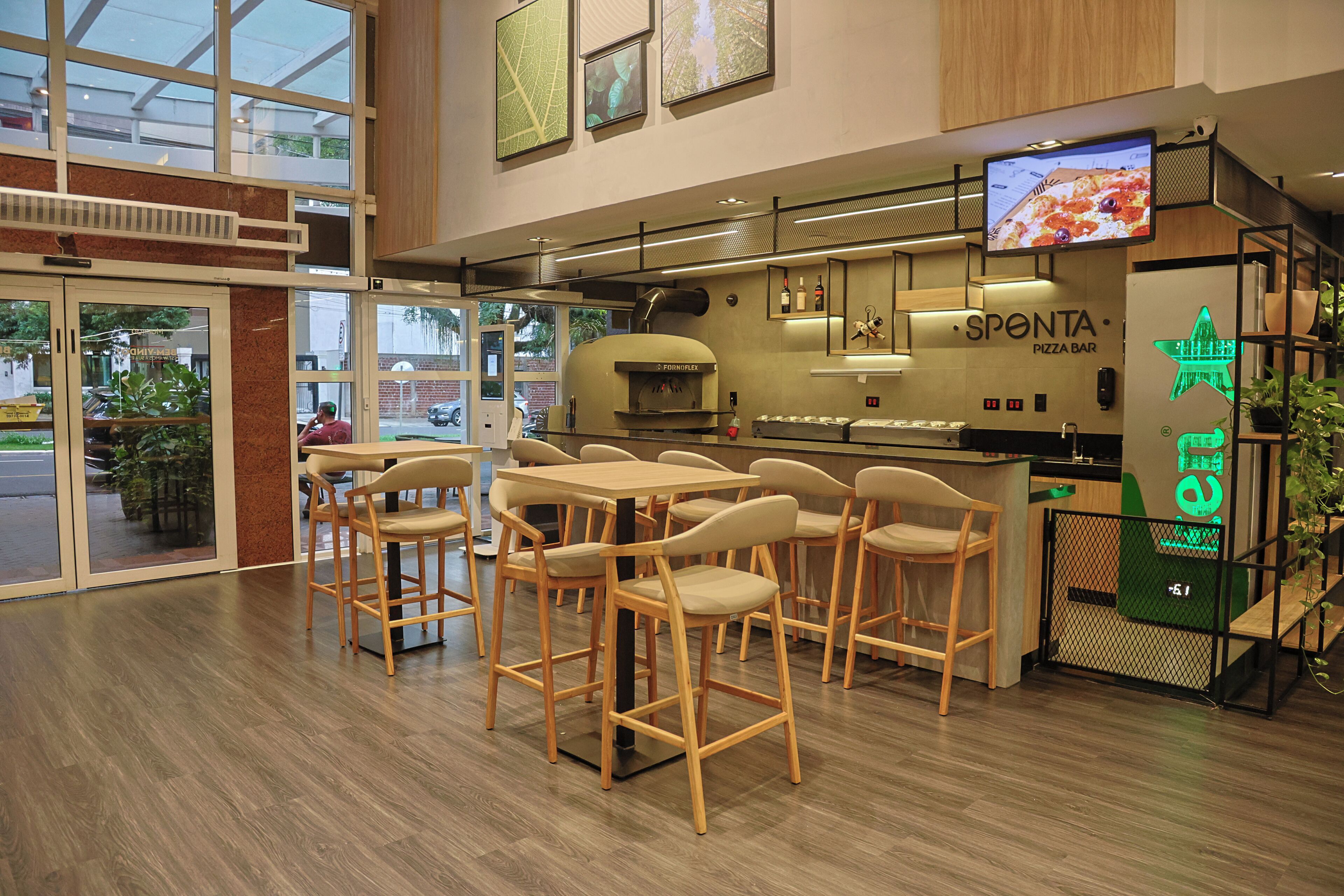 A modern pizza bar interior with wooden chairs and tables, a pizza oven, and a digital menu display.