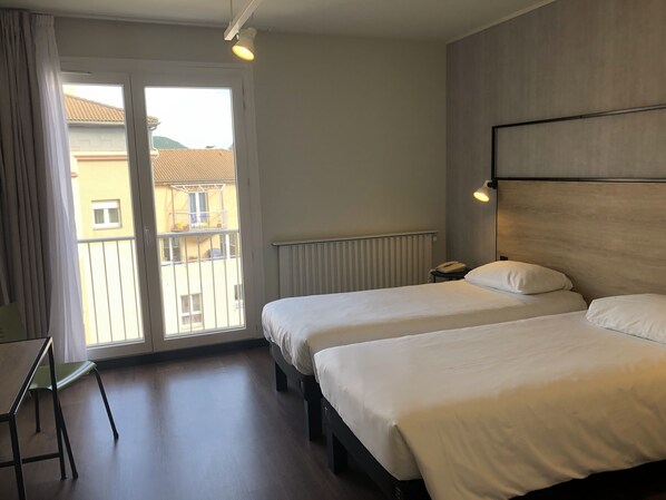 A hotel room with two beds, a desk, and a chair. There is a window with a view of buildings outside.