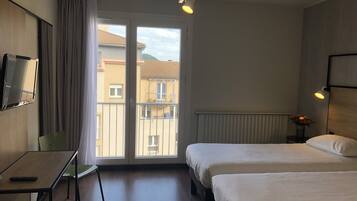 A hotel room with two beds, a desk, a TV, and a balcony with a view of buildings.