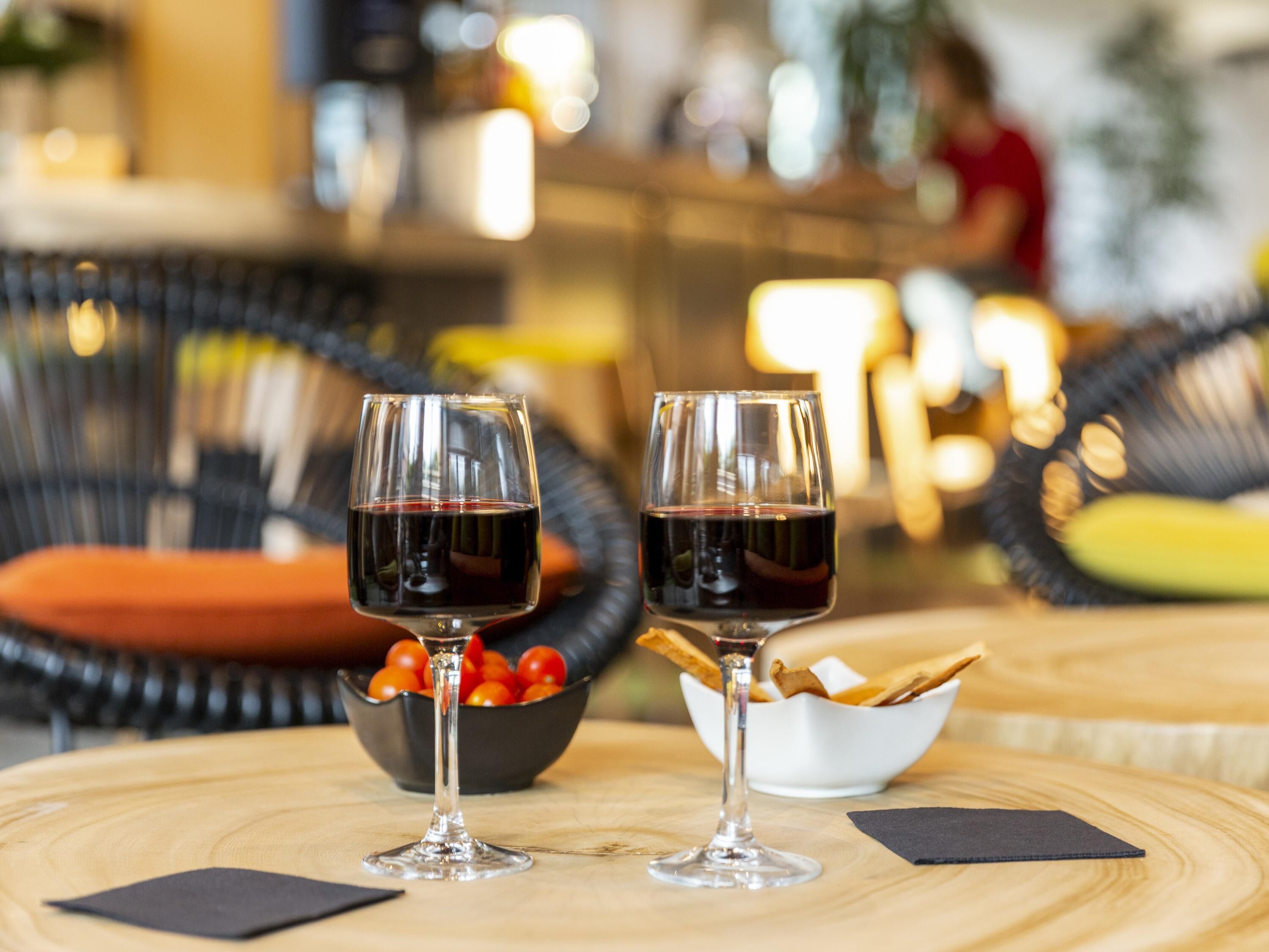 Two wine glasses on a wooden table with a bowl of cherry tomatoes and a bowl of crackers.