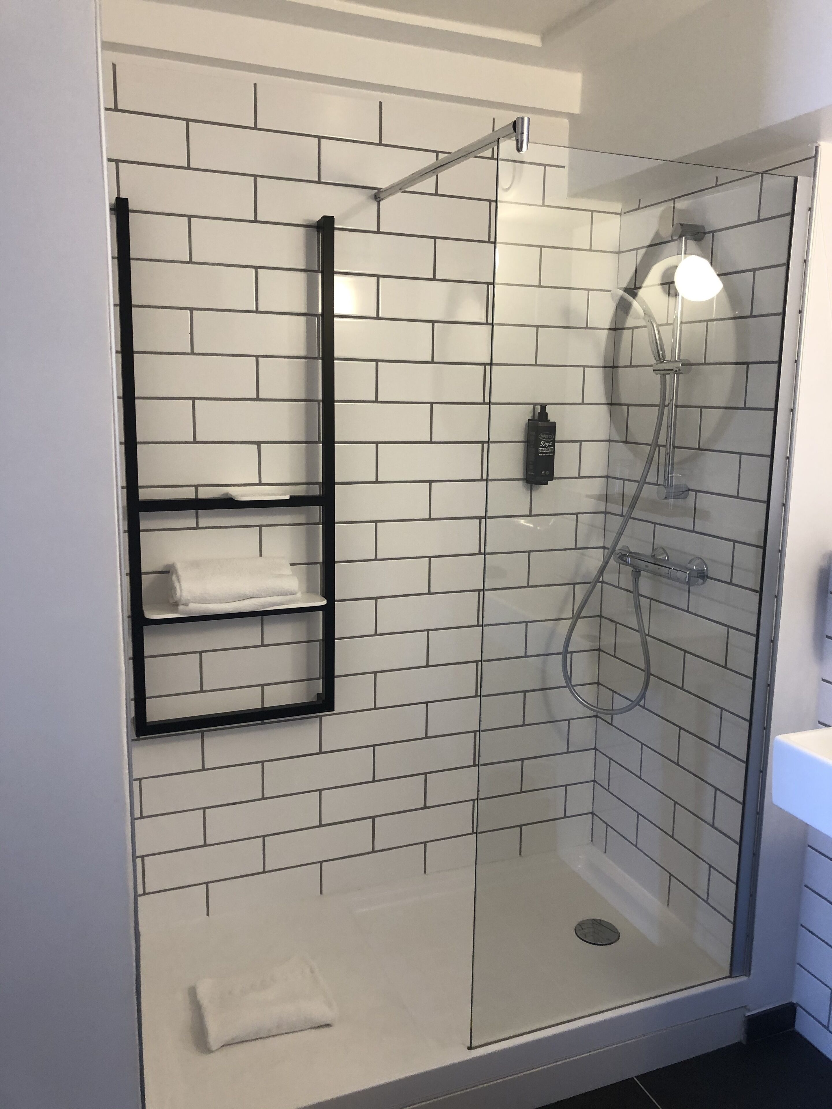 Una ducha moderna con puerta de vidrio, paredes con azulejos blancos y un perchero negro.