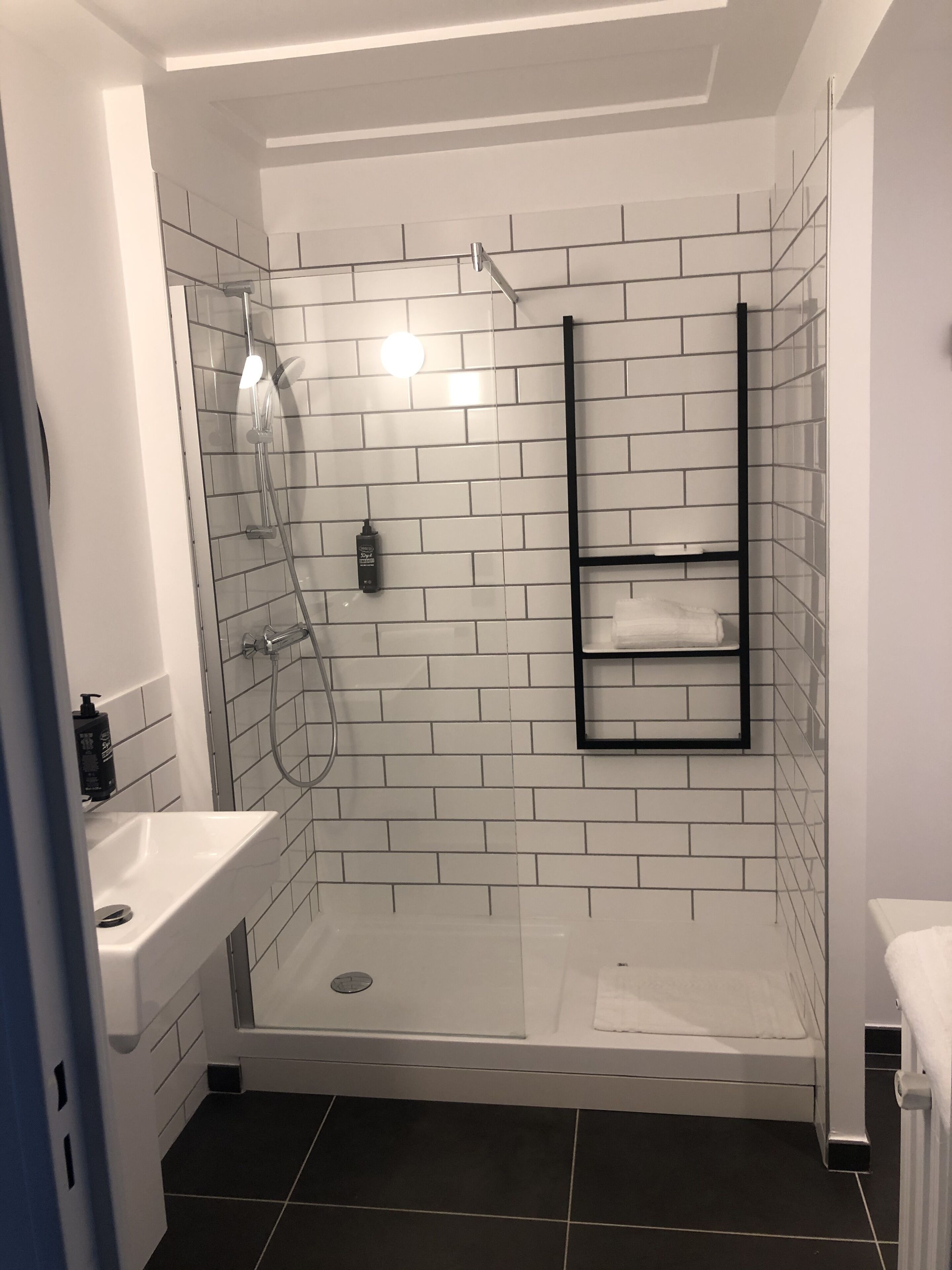 Un baño moderno con ducha envuelta en vidrio, paredes de azulejos blancos y estante con marco negro.