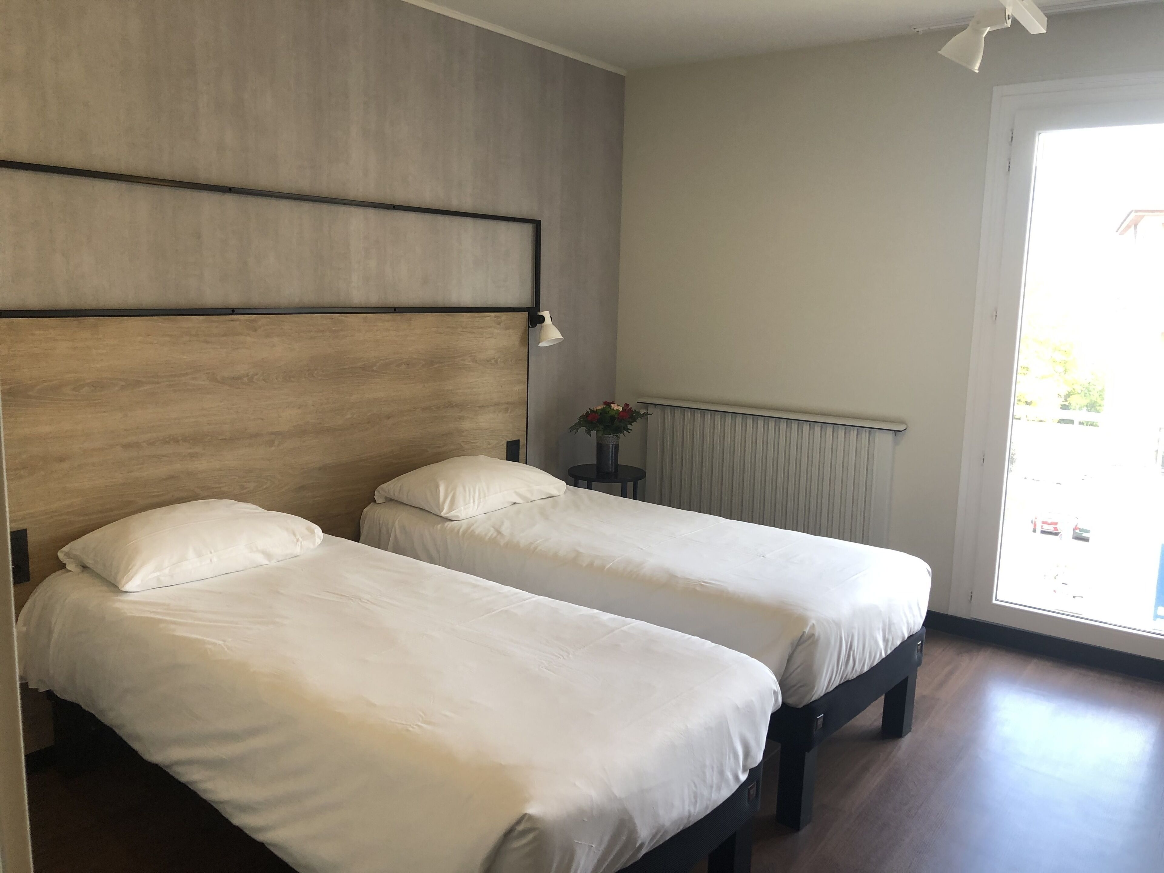 A room with two single beds, a wooden headboard, a small table with a vase of flowers, and a window with a view of the street.