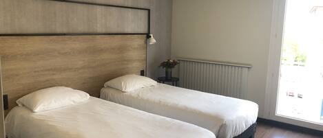 A room with two single beds, a wooden headboard, a small table with a vase of flowers, and a window with a view of the street.