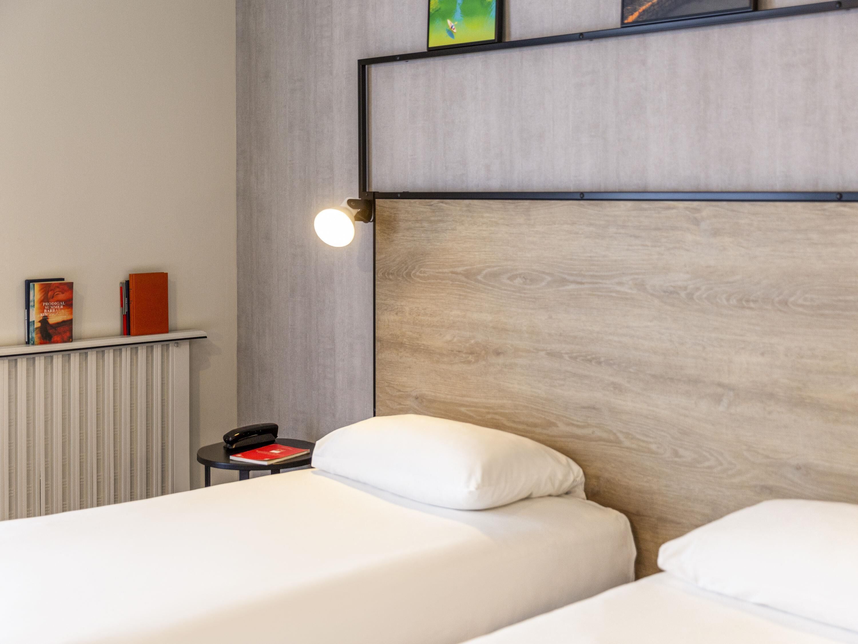 A modern hotel room with a wooden headboard, two white beds, a bedside table with a phone and a red book, and a radiator.