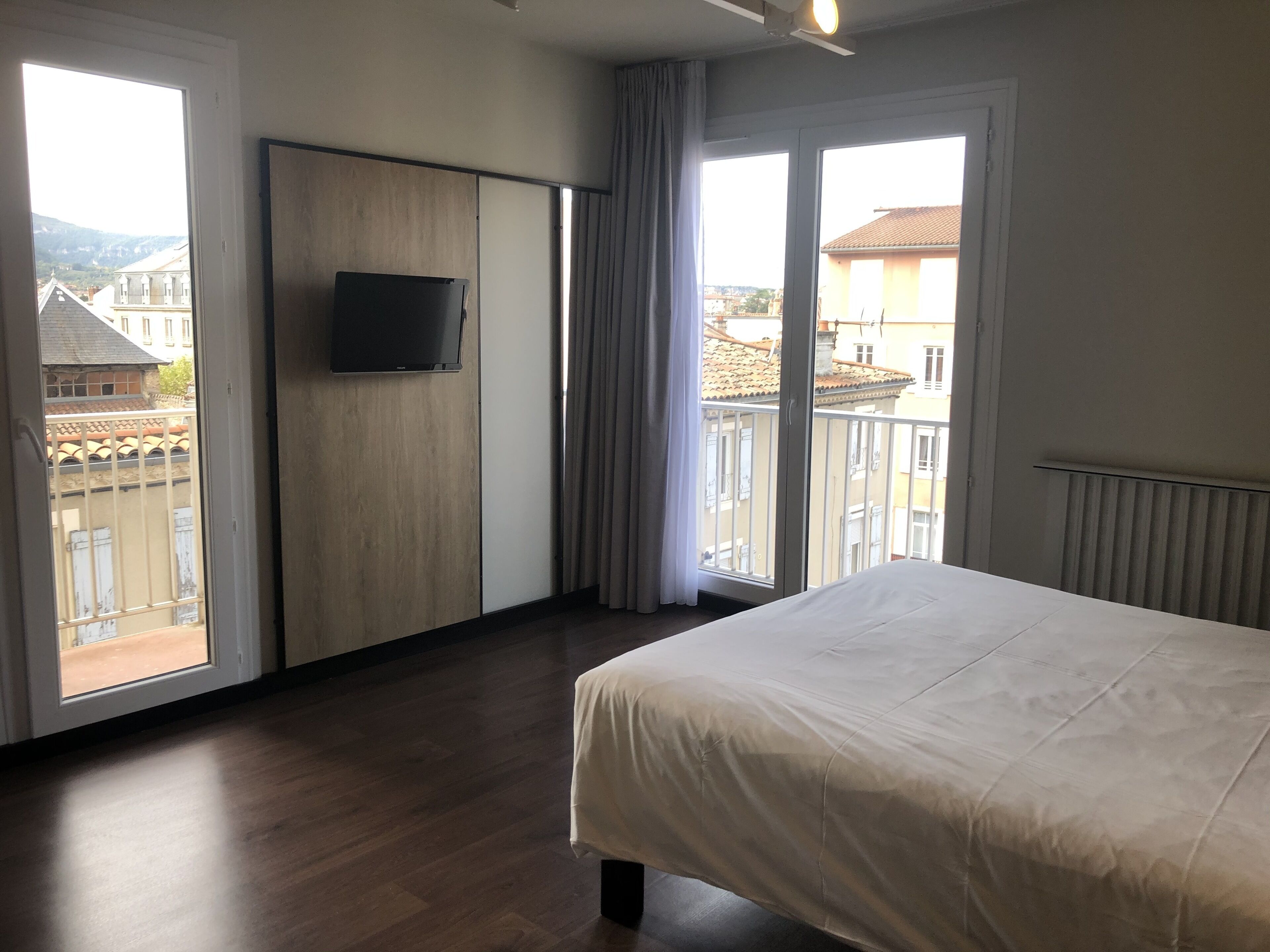 A hotel room with a bed, a TV mounted on the wall, and large windows offering a city view.