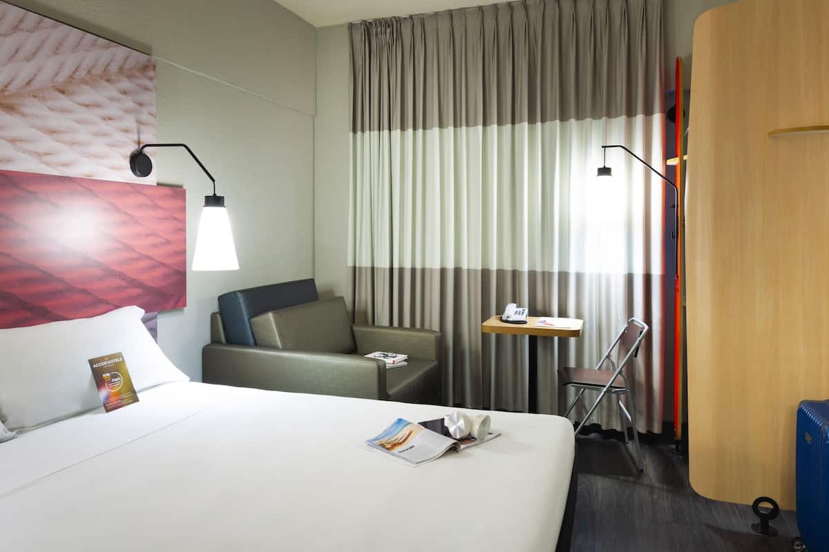 A hotel room with a bed, a desk, a chair, a lamp, and a curtain.