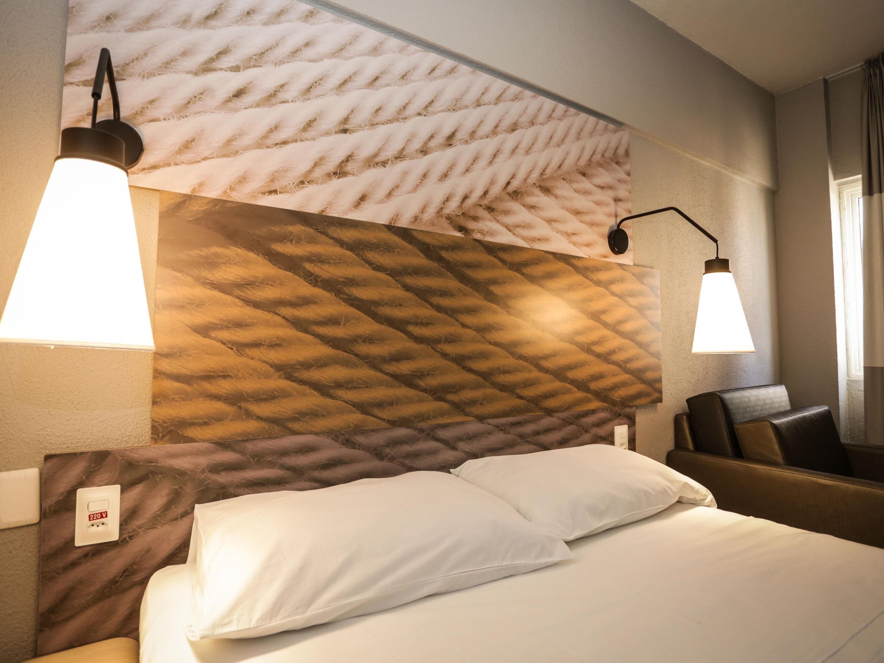 A hotel room with a bed, two bedside lamps, a headboard with a textured pattern, a chair, and a wall socket.